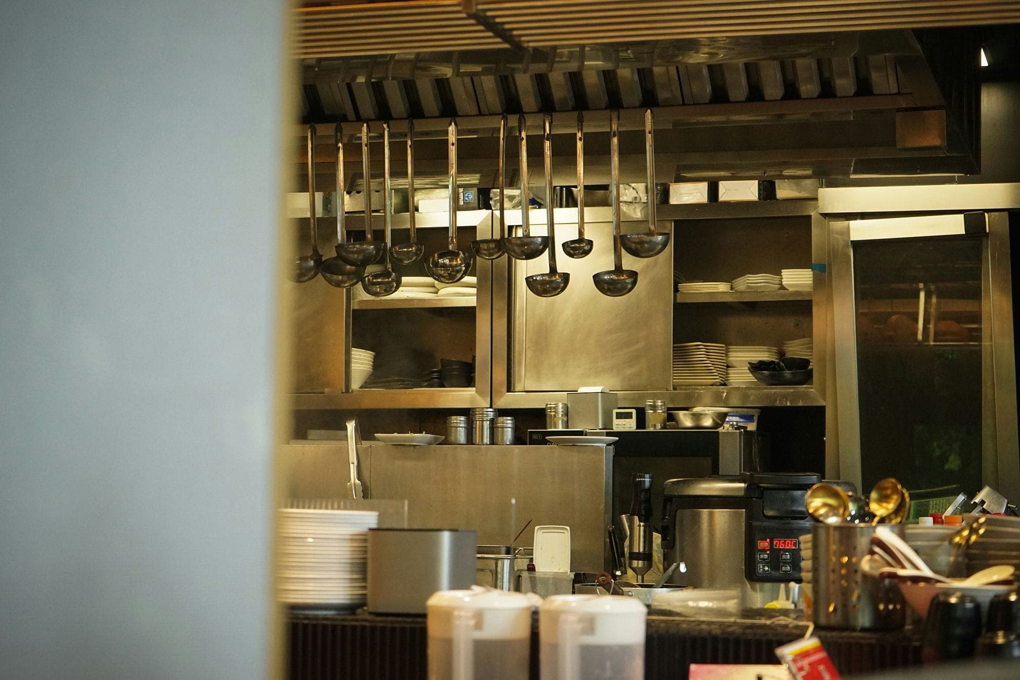 Photograph of a restaurant kitchen, focusing on a wall of cabinets full of dish ware. In the foreground, a collection of ladles hang from a rack and a variety of utensils, dish ware, and pots are covering the counter.