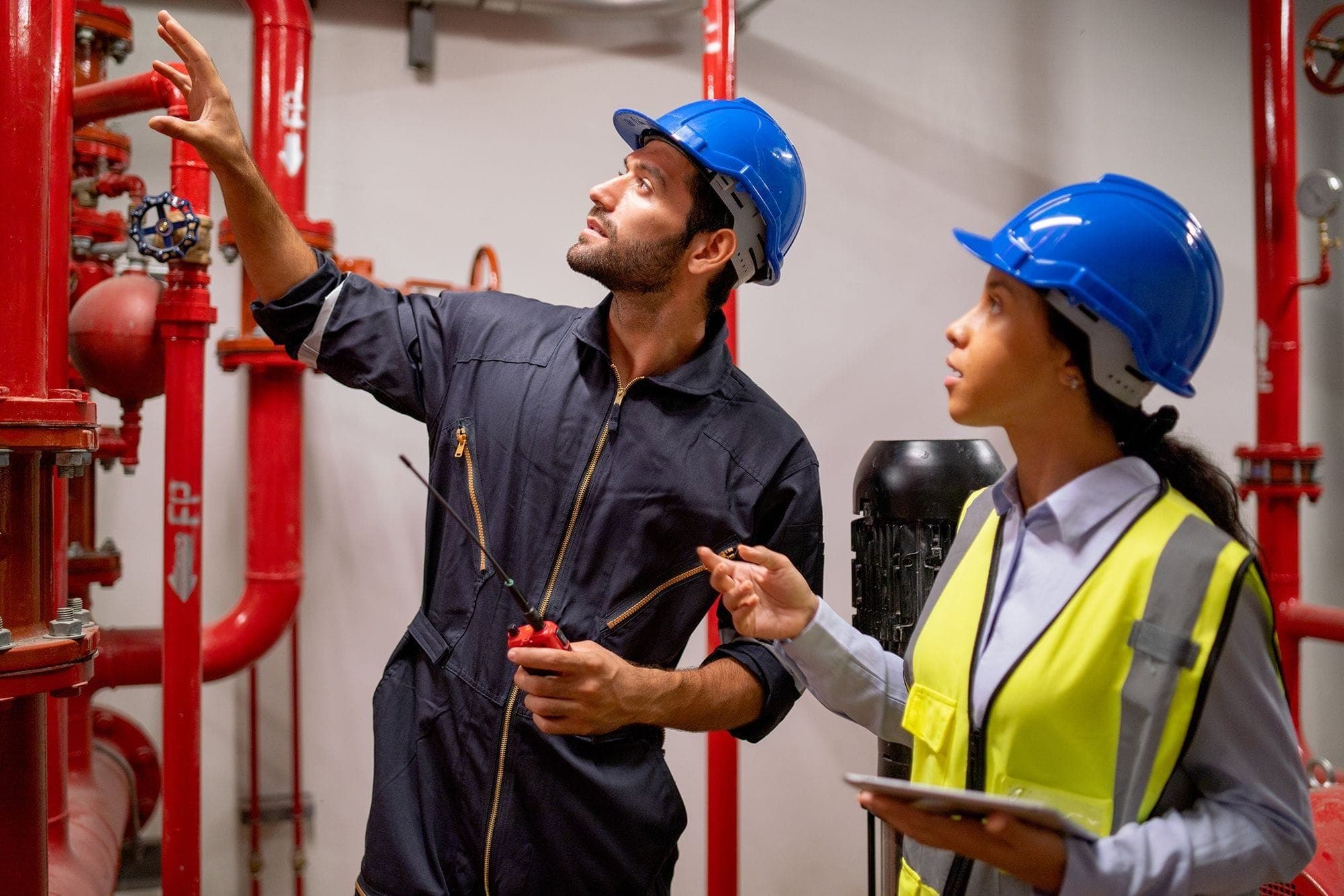 Photograph of two fire safety inspectors, with one pointing at a piece of equipment on the wall while the other holds a clipboard. They appear to be discussing the item at which the first is pointing. Both are wearing protective gear.
