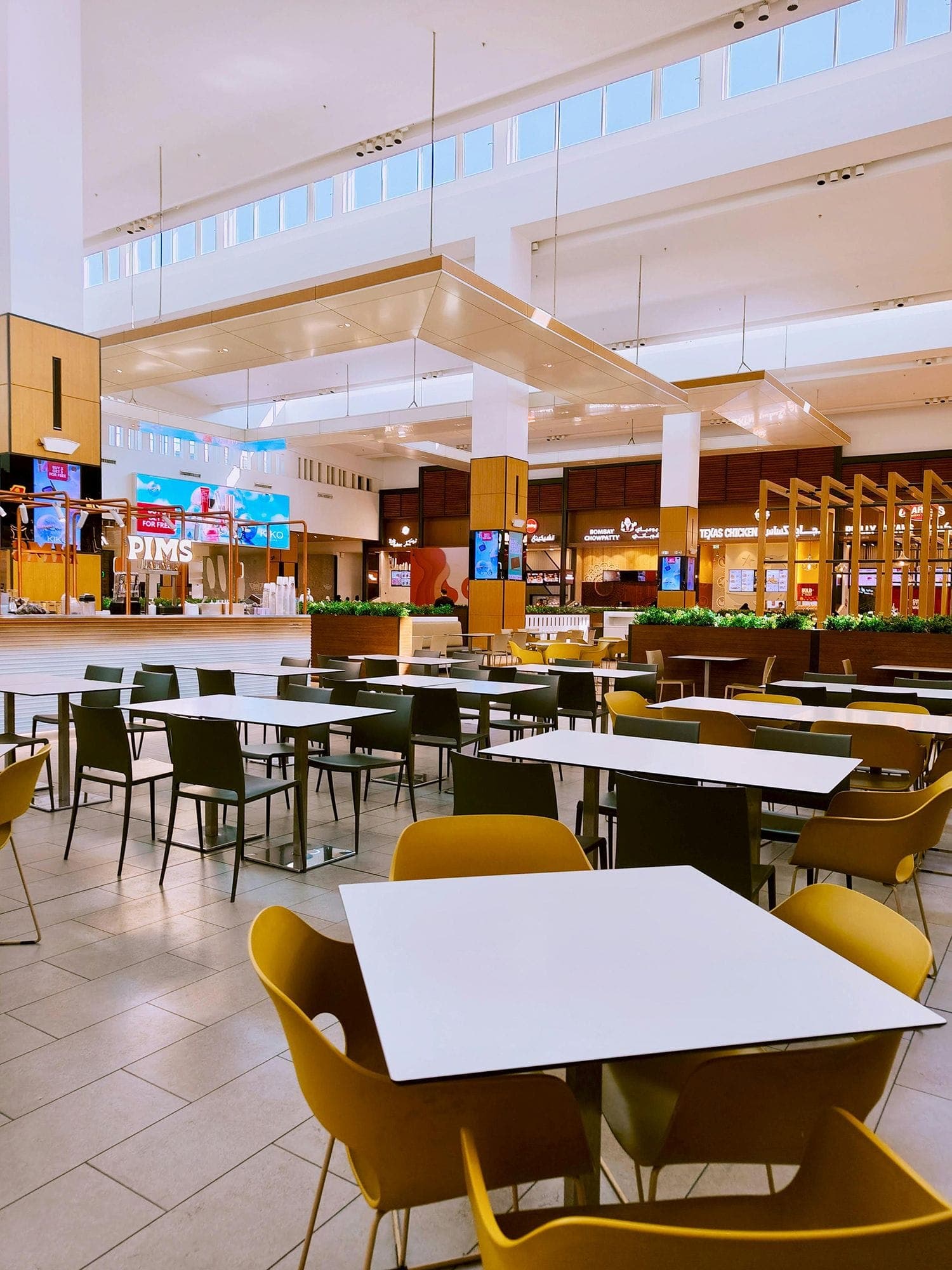 Photograph of a mall food court; many tables are visible in the foreground, with multiple fast food storefronts in the background.