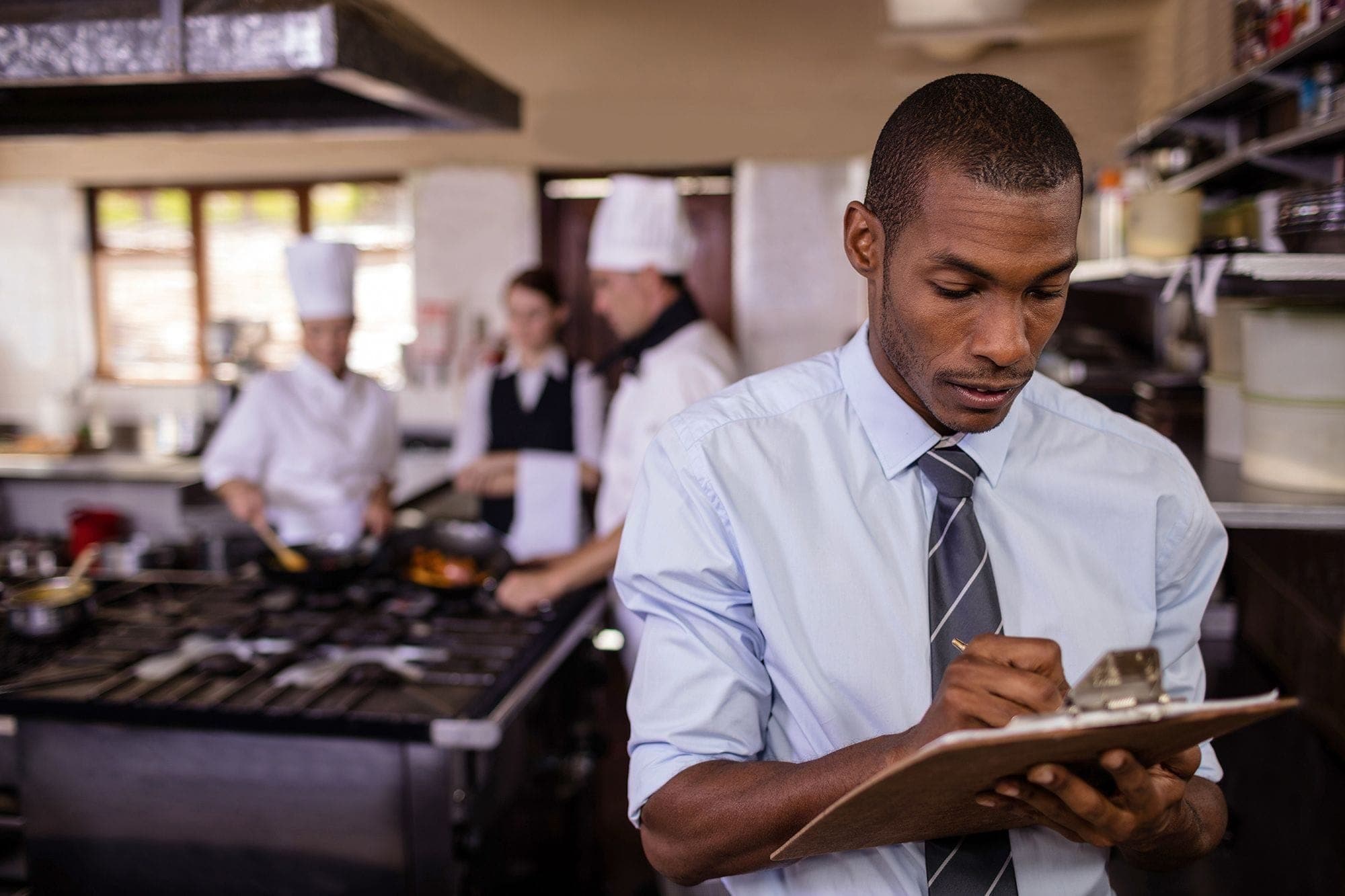 Photograph of an inspector standing in a commercial kitchen, taking notes on a clipboard. Behind him, chefs and other restaurant employees are gathered in the background.