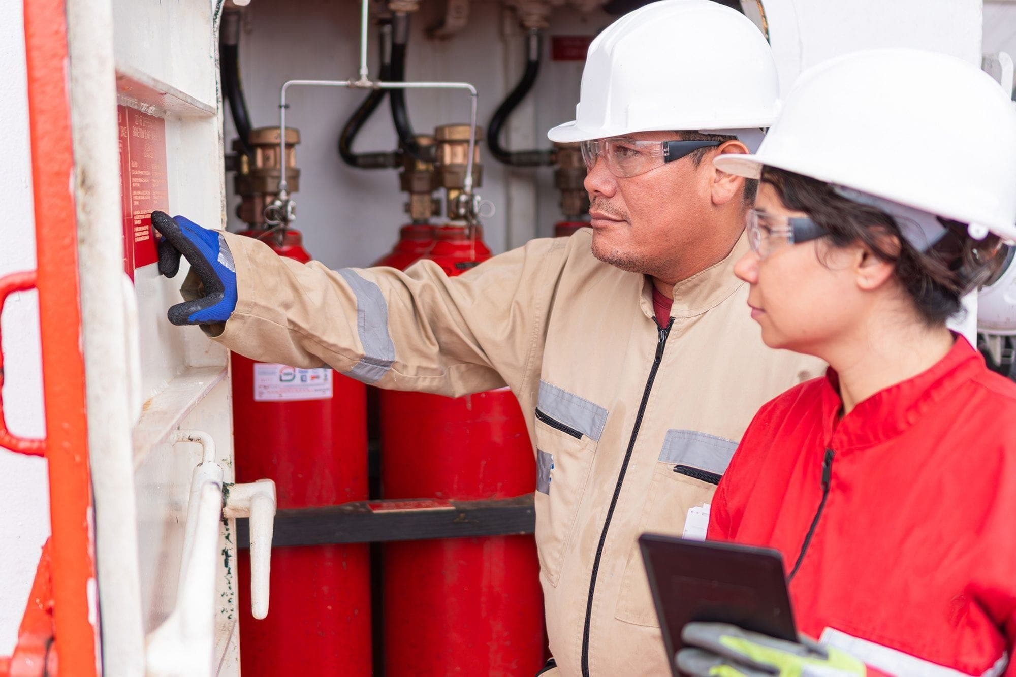 Photograph of two fire safety inspectors, with one pointing at a piece of notation on the wall while the other holds a clipboard. Both are wearing protective gear.
