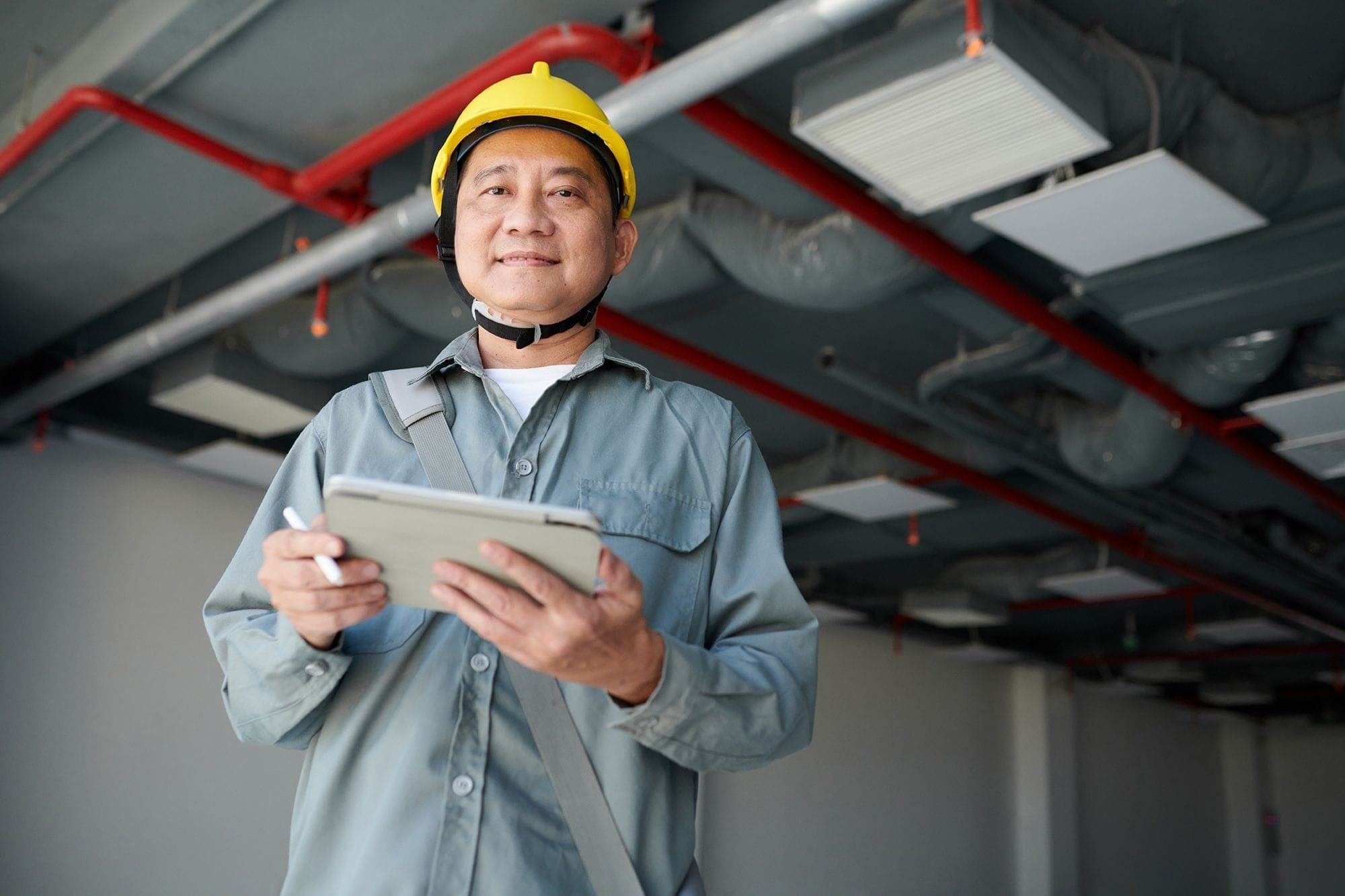 Photograph of a fire safety inspector, with a visible fire safety sprinkler system on the ceiling above him, holding a clipboard and smiling at the camera.