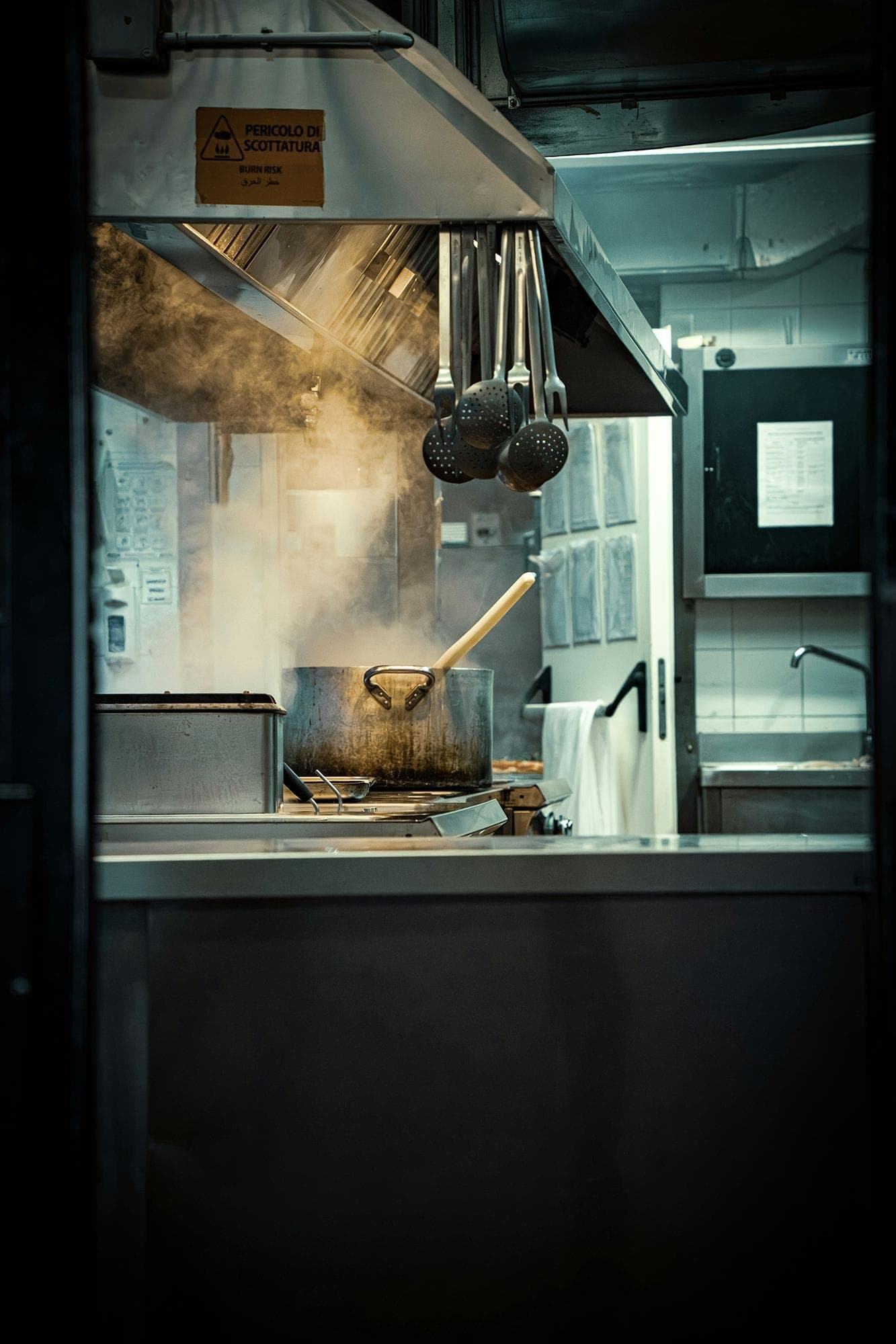 Photograph of a restaurant kitchen, focusing on a pot over a heated stove. Ladles hang from above and a sink is visible in the background.